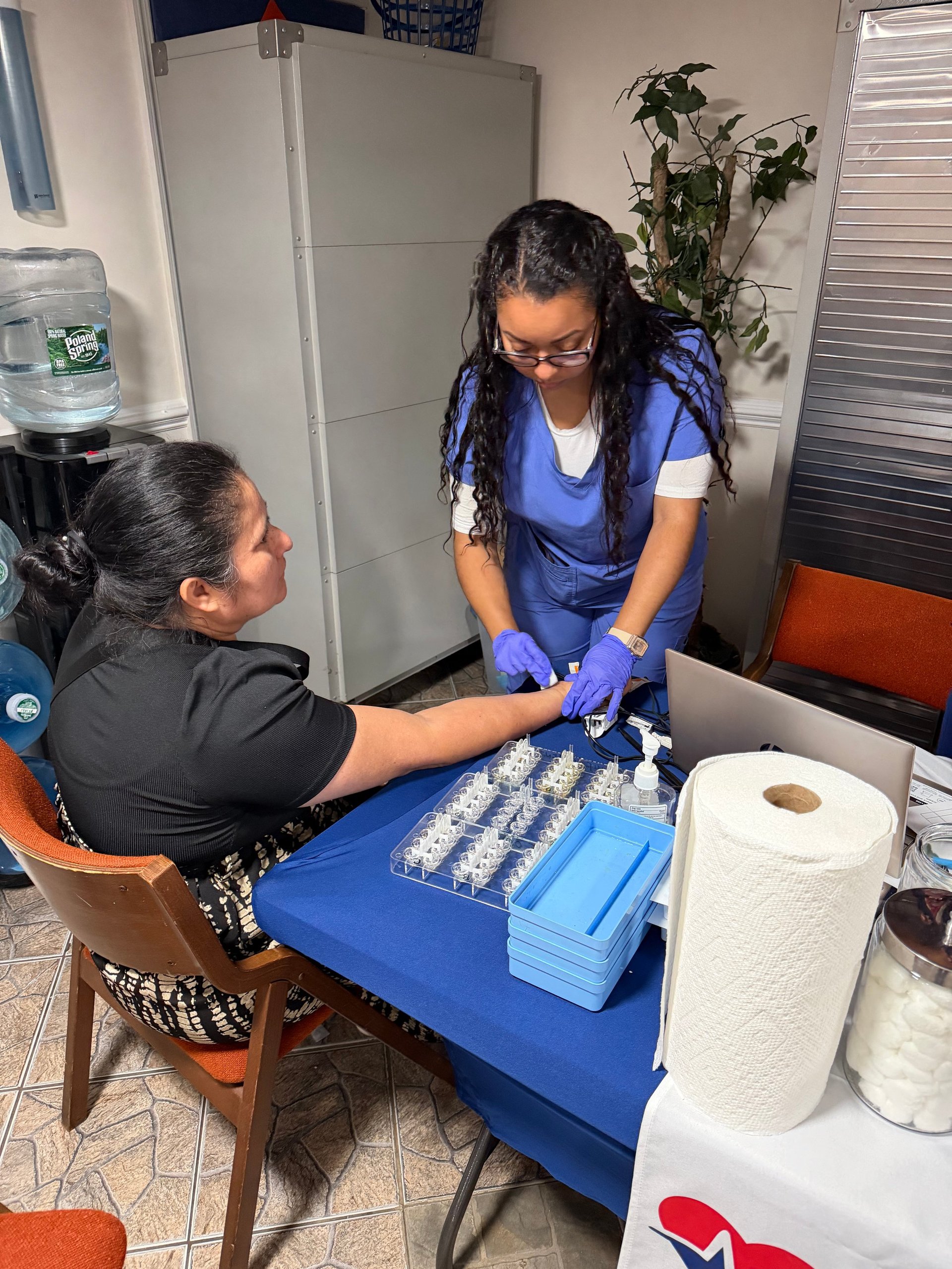 Healthcare worker in blue gloves and scrubs organizing medication samples on a blue table with patient seated nearby