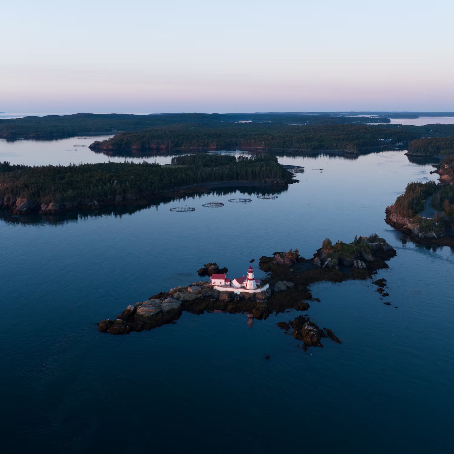 Aerial view of a red and white lighthouse on a rocky island surrounded by calm blue water and forested shoreline at dusk