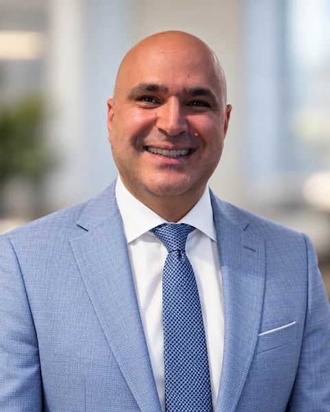 Professional headshot of a bald man in a light blue suit and blue patterned tie, smiling at the camera with city buildings in the background