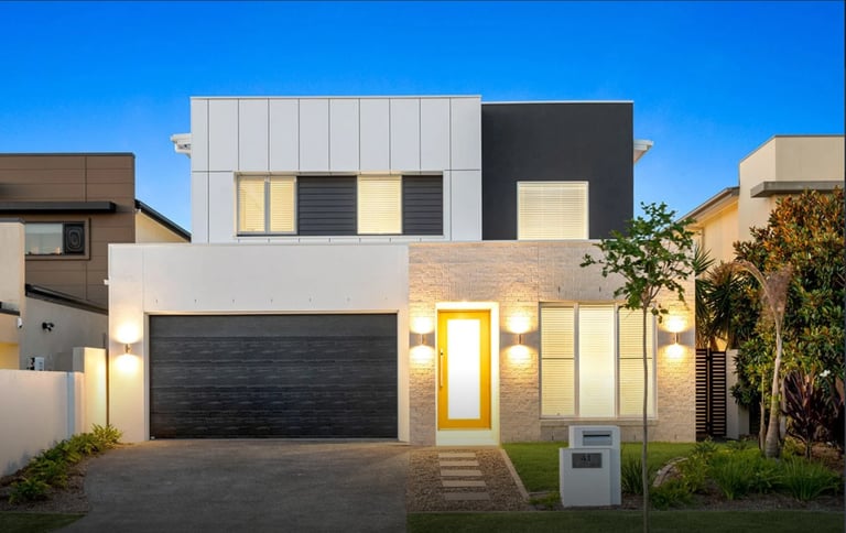 Modern two-story house with white brick facade, dark garage door, and illuminated entrance at dusk