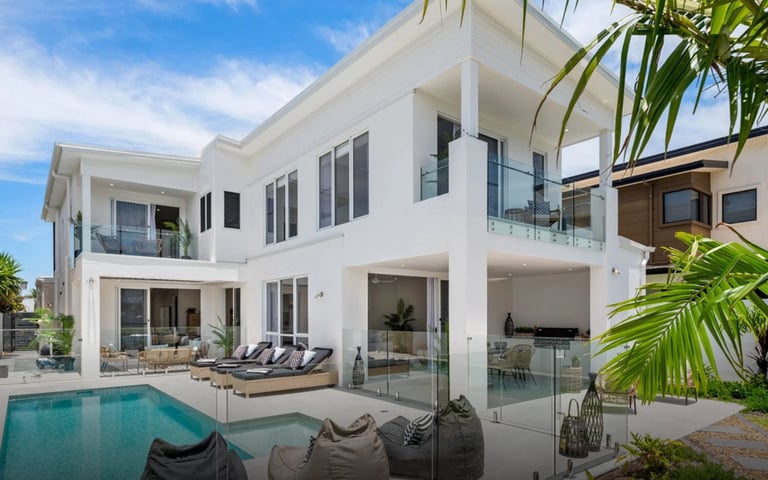 Modern white two-story house with pool, lounge chairs, and tropical plants in sunny backyard setting
