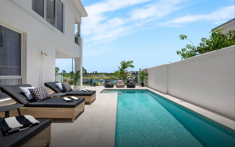 Modern home with rectangular swimming pool, lounge chairs, white patio deck, and tropical landscaping under blue sky