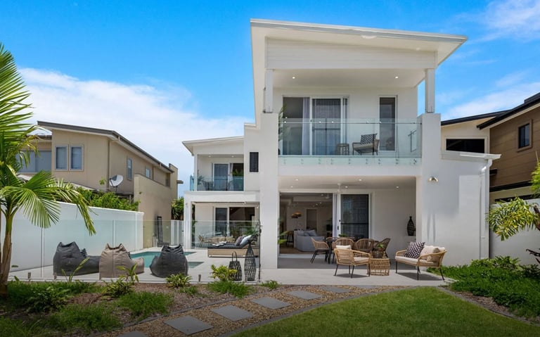 Modern three-story white house with large balcony, patio furniture, and landscaped yard on sunny day