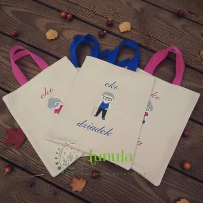 Three custom tote bags with pink and blue handles on wooden surface with scattered beads and autumn leaves