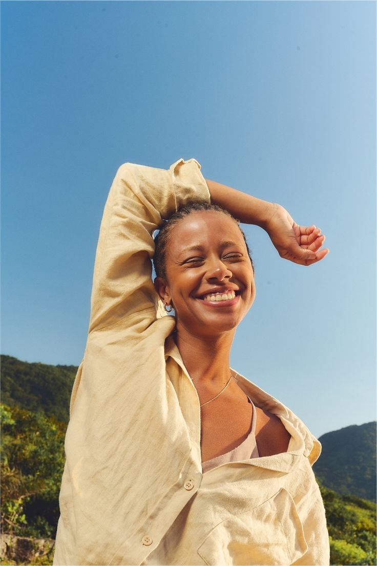 Smiling woman in cream cardigan with hand behind head, outdoors against blue sky and green hills