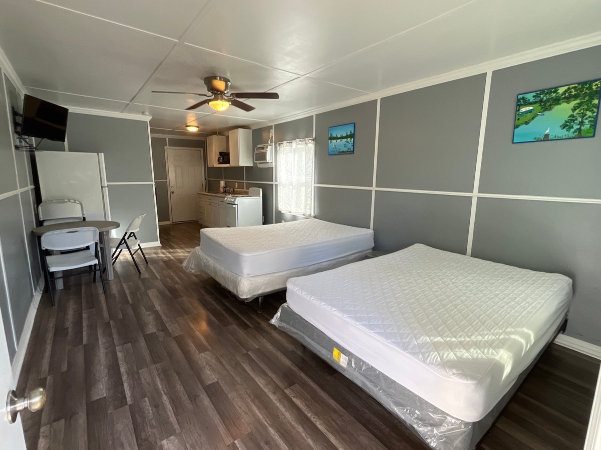 Cabin bedroom with two white beds, gray walls, ceiling fan, and kitchenette in background