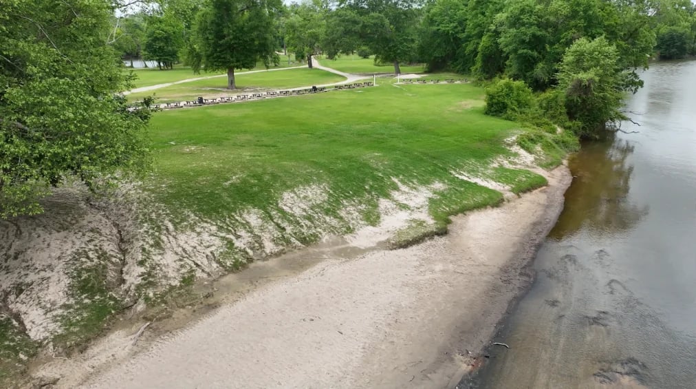 Aerial view of a grassy park with sandy riverbank and water on the right side, lined with green trees