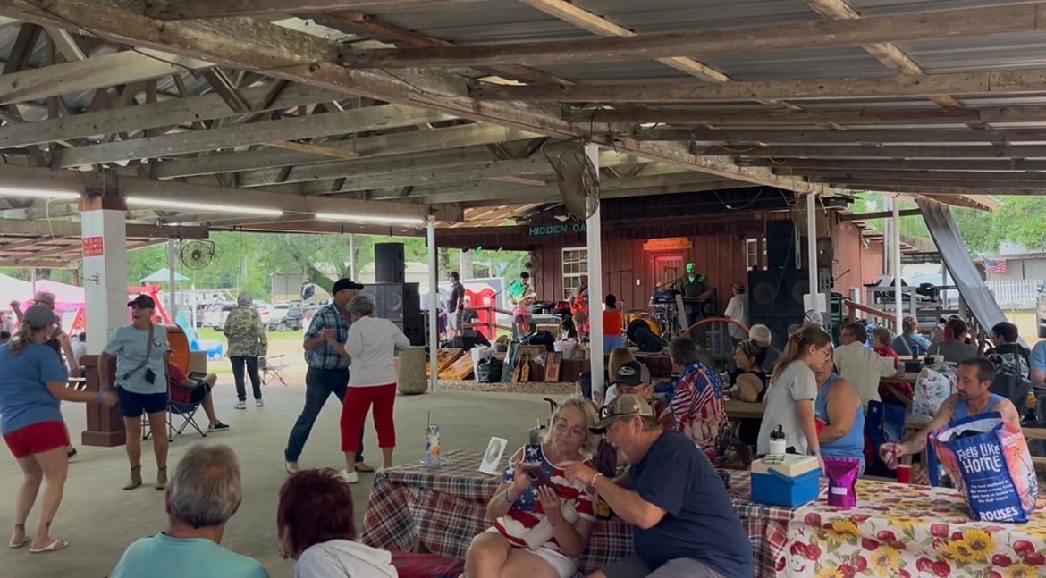 Outdoor community event or market under a covered pavilion with vendors and shoppers browsing tables and walking around