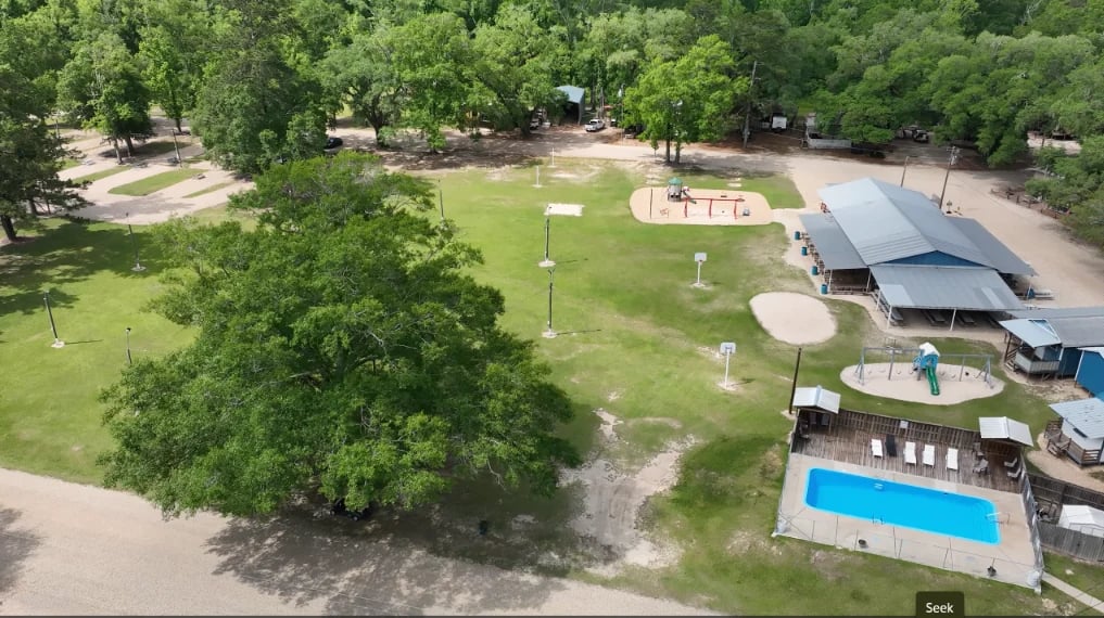 Aerial view of a park with green lawn, playground equipment, pavilion, and swimming pool surrounded by trees