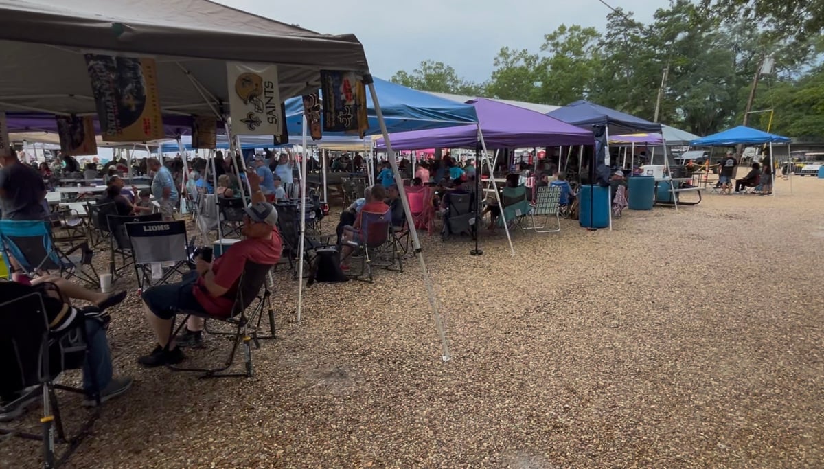 Outdoor vendor market with multiple colorful canopy tents and shoppers on a dirt lot with trees in background