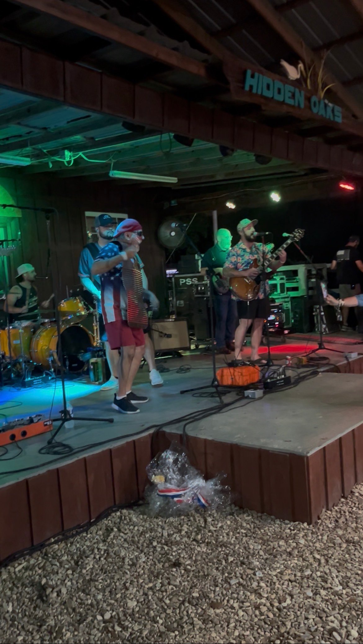 Band performing on stage at Hidden Oaks venue with green neon lighting, drums, and musicians with instruments