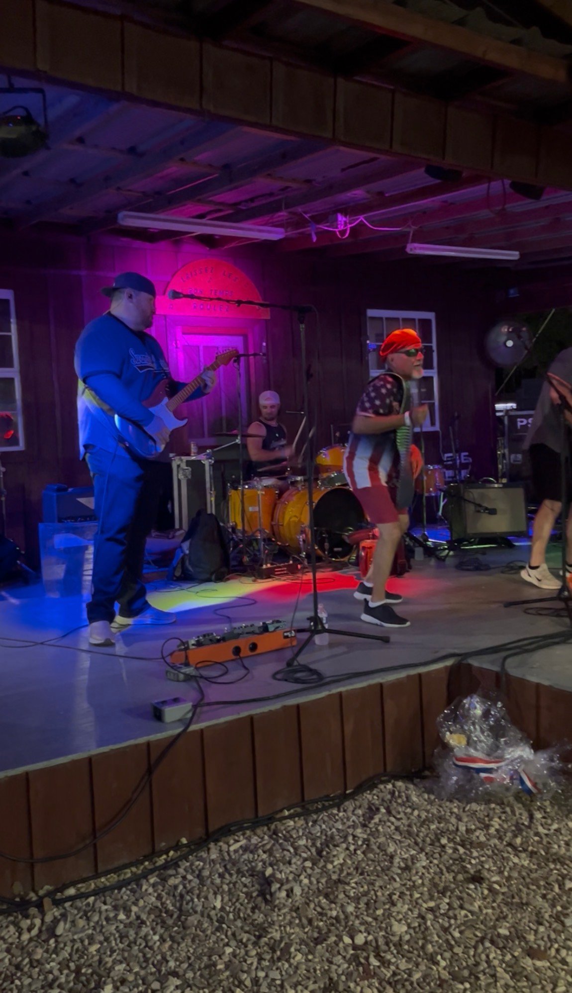 Live band performing on stage with purple neon lighting, featuring guitarist in blue shirt, drummer, and vocalist in orange cap