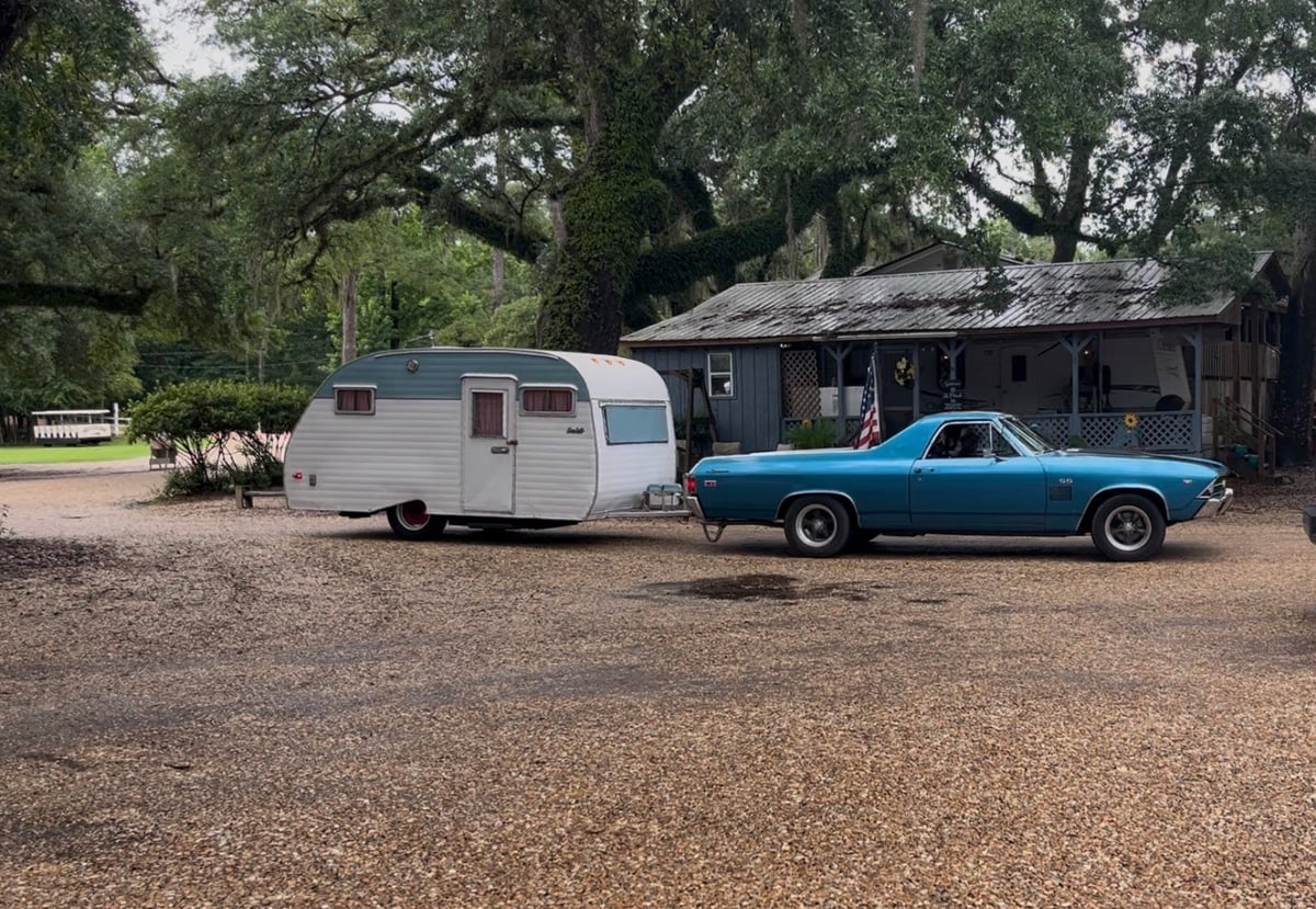 A blue vintage car parked next to a white travel trailer in front of a rustic cabin, surrounded by large oak trees.