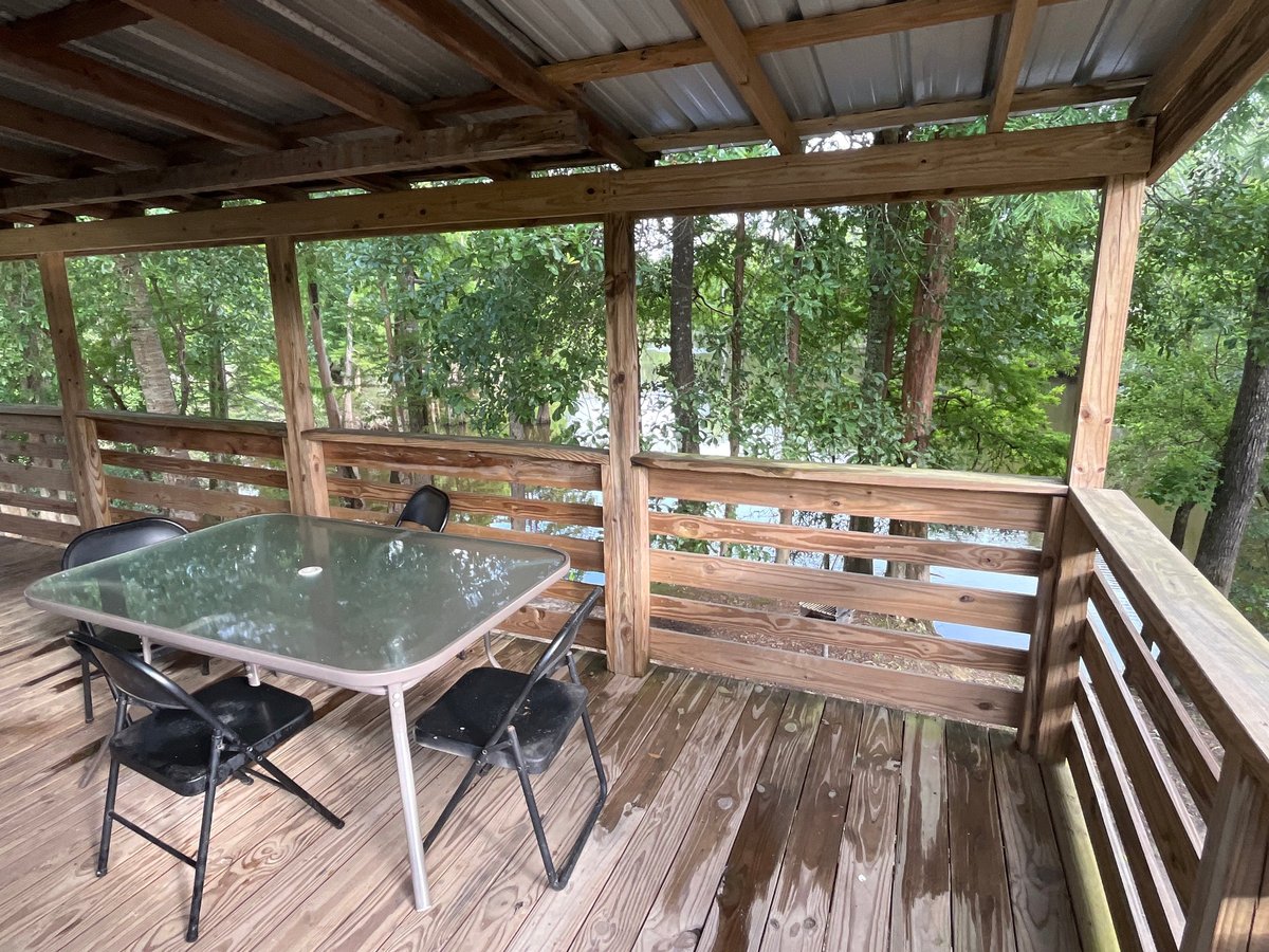 Covered wooden deck with glass table and black chairs surrounded by trees
