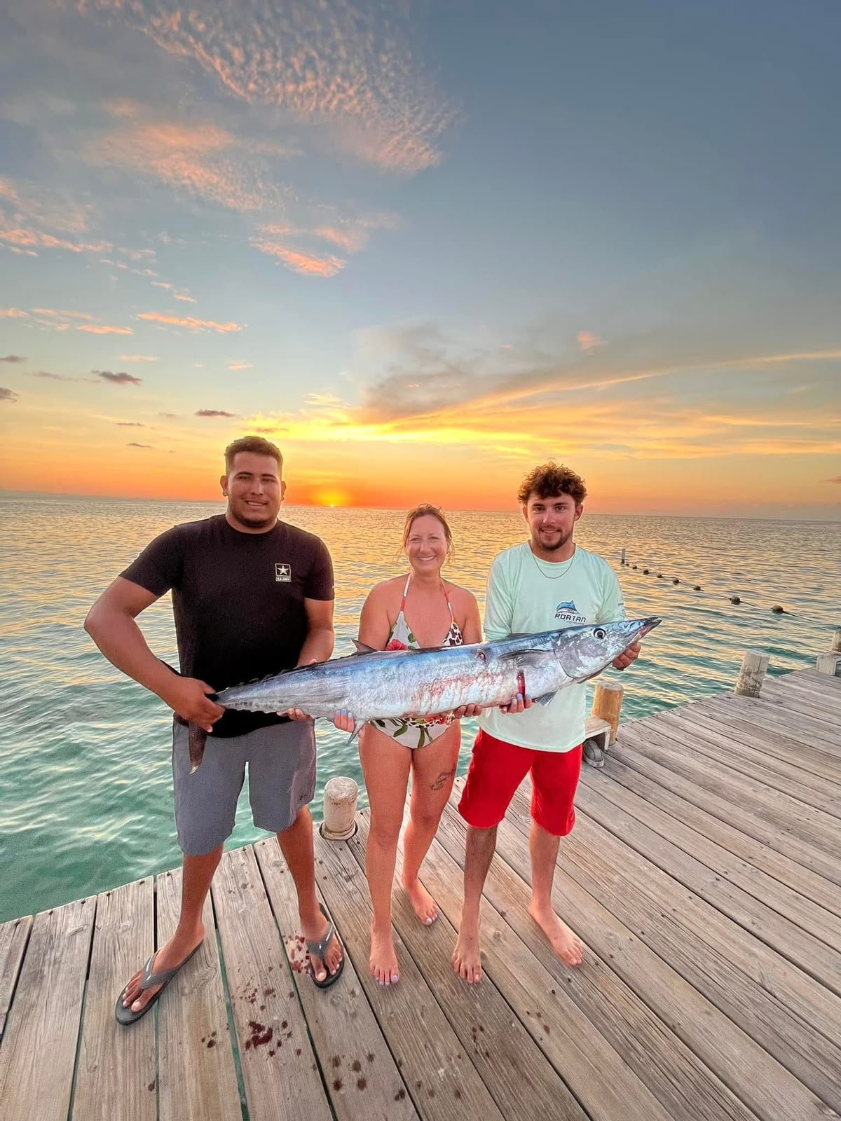 Three guests holding a large king mackerel on a wooden dock at sunset in Roatán, Honduras after a successful fishing charter with Gustavo Pavon Tours