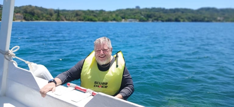 Elderly man in yellow life vest smiling while in water next to a boat, with forested hills in the background