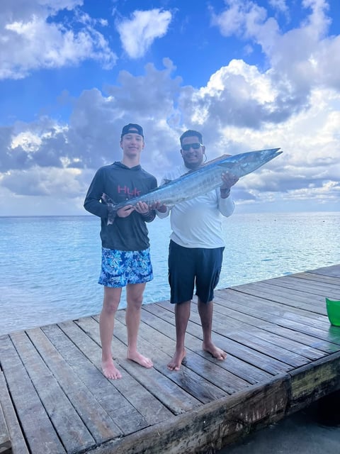 Two men on a wooden dock holding a large blue barracuda with ocean and clouds in the background after a Roatán fishing charter