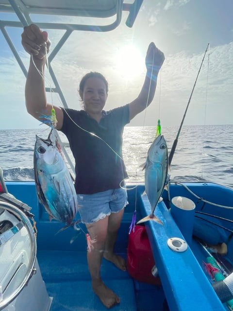 Angler on a Roatán fishing boat holding up two freshly caught blackfin tuna with arms raised, Caribbean ocean in the background