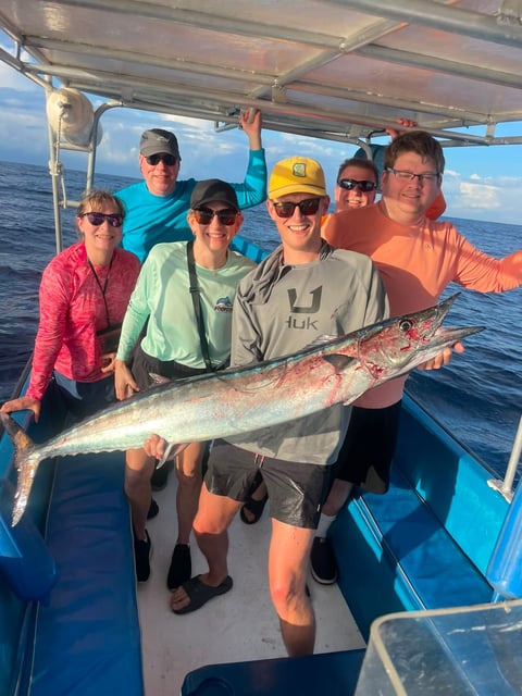 Group of six people on a Roatán fishing charter boat holding a large wahoo with Caribbean ocean and blue sky in the background