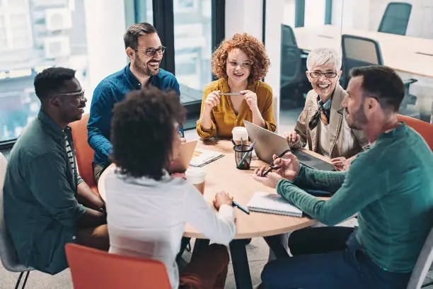 Diverse team of six professionals having a discussion around a wooden table in a modern office setting with large windows