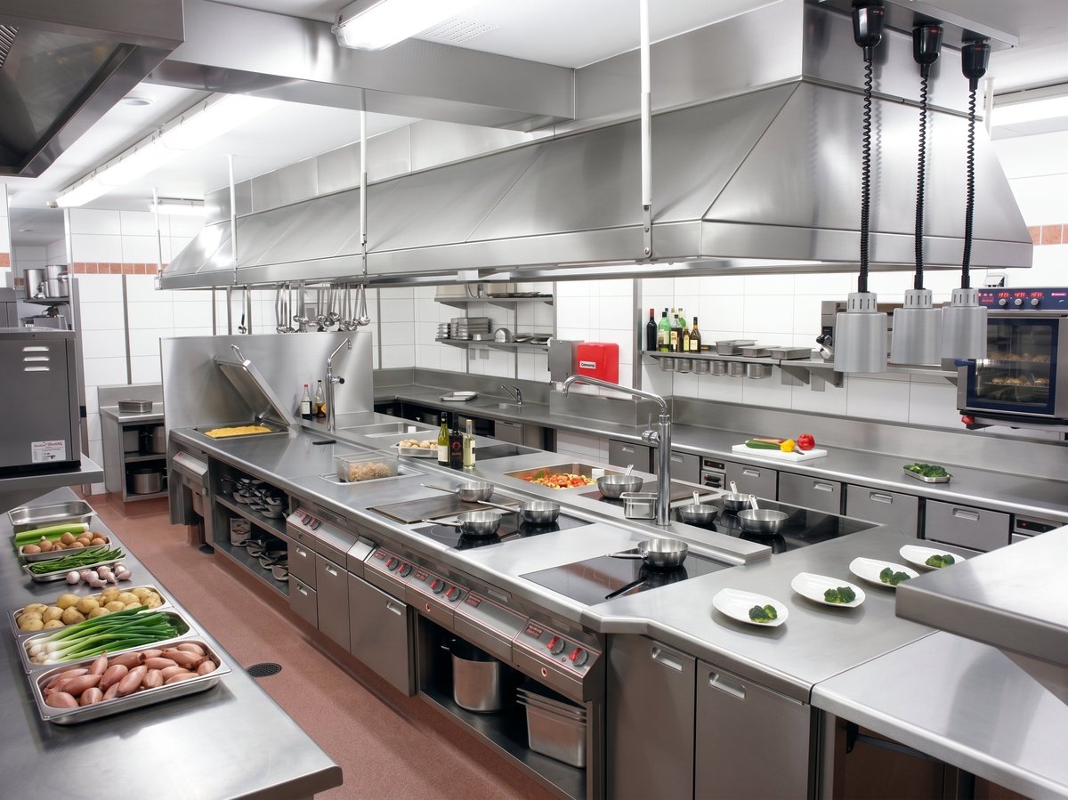 Professional stainless steel commercial kitchen with overhead ventilation, cooking stations, and prep counters lined with ingredients