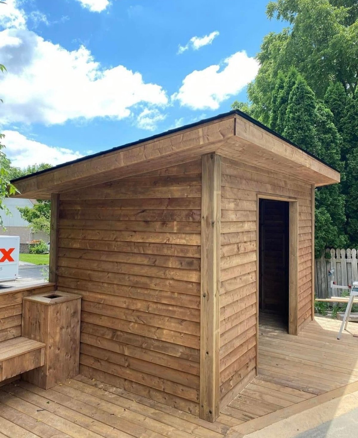 Wooden garden shed with horizontal siding on a deck, surrounded by green trees and blue sky