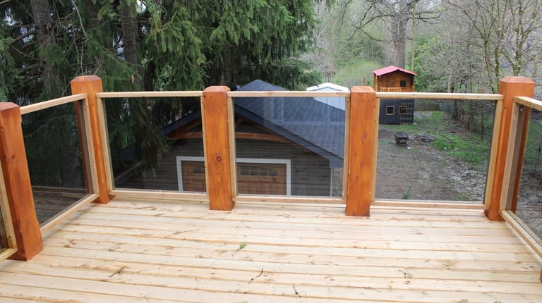Wooden deck with glass railings and wooden posts overlooking a wooded backyard with structures visible in the distance