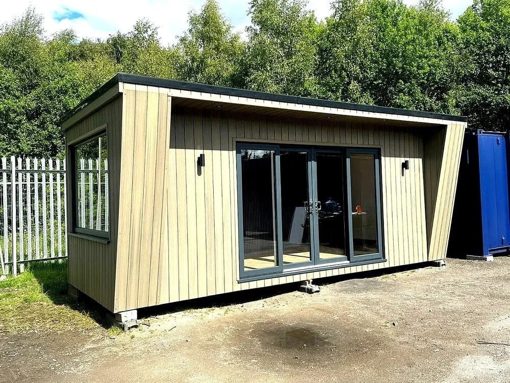 Modern cream and black modular garden office cabin with large sliding glass doors and white fence, set against trees and green lawn