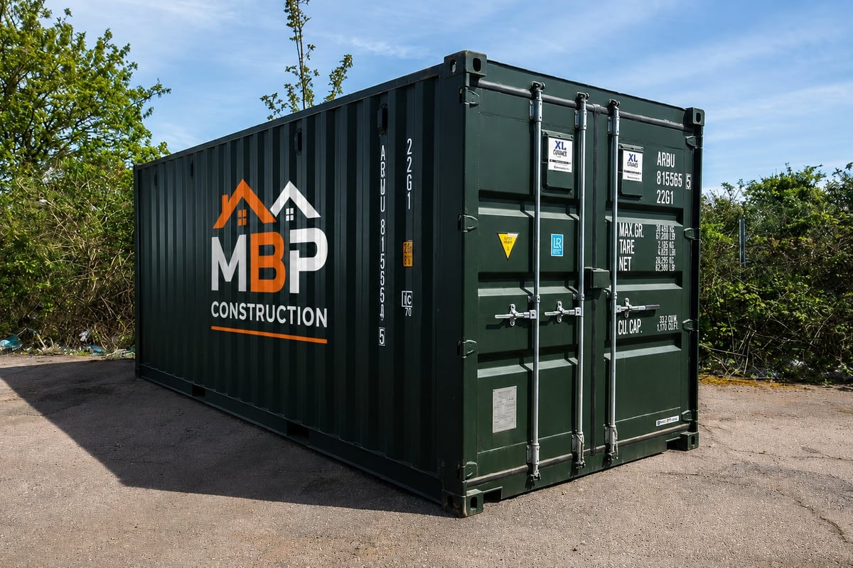A green and black shipping container with MBP Construction logo and branding on the side, positioned outdoors with trees in the background.