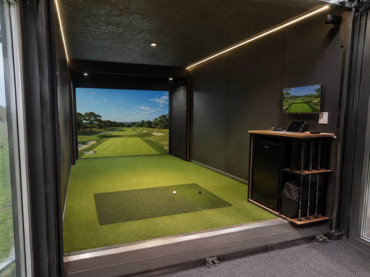 Indoor golf simulator room with green artificial turf, hitting mat, screen displaying golf course, and equipment shelving on right wall