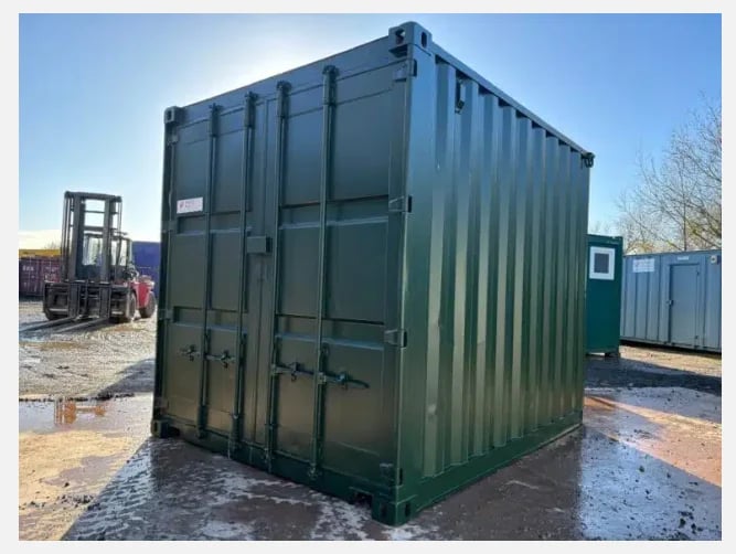 Green shipping container on wet ground with forklift and other containers visible in background on industrial site