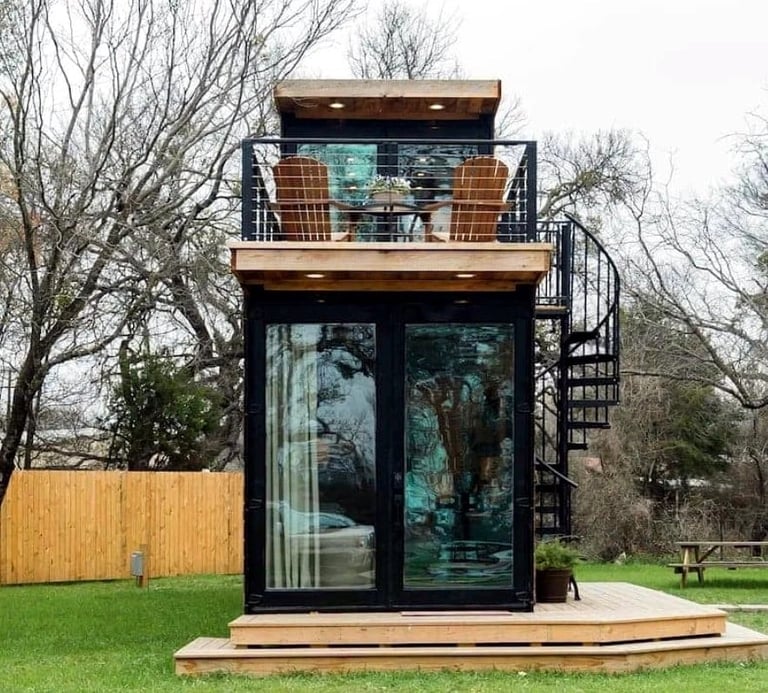 Modern two-story shipping container tiny house with black metal frame, large windows, and rooftop terrace in a grassy field surrounded by bare trees