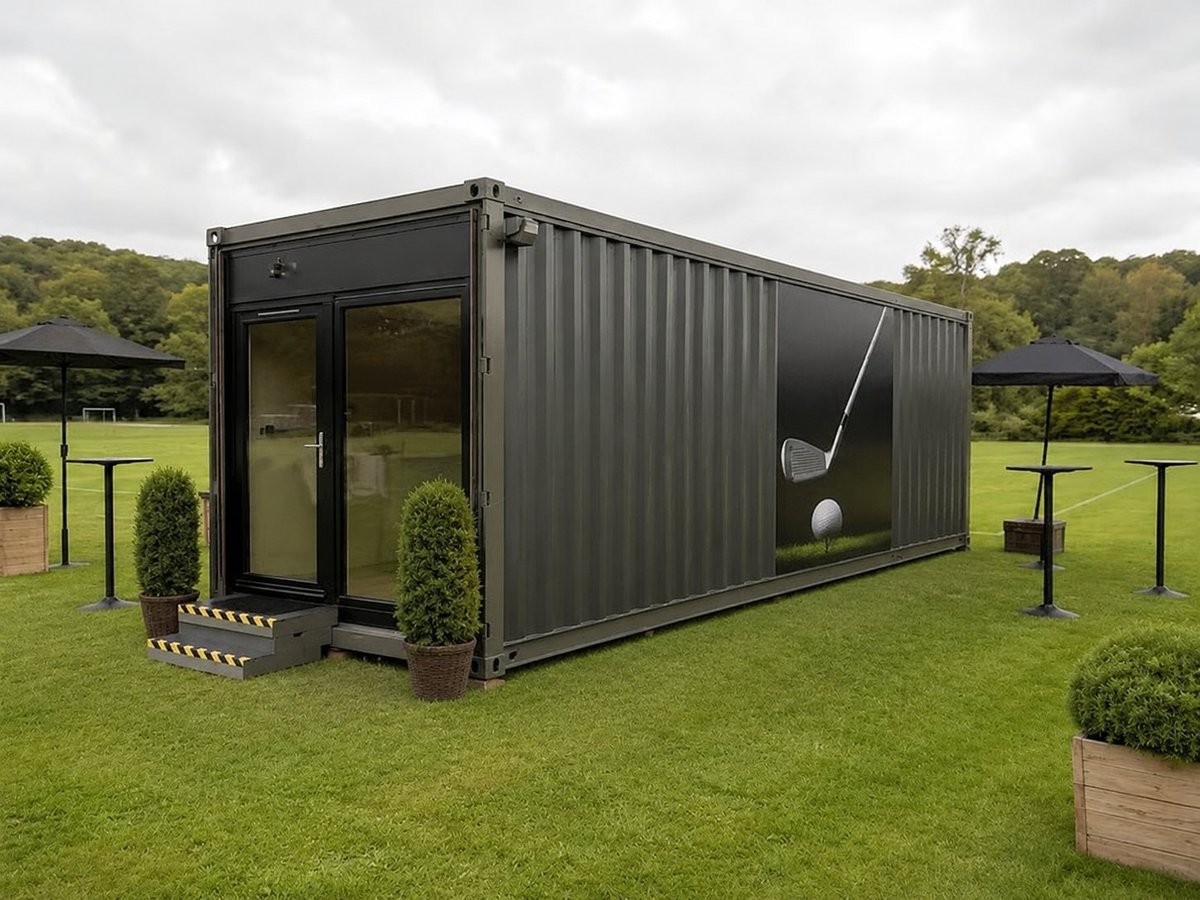 Modern black shipping container conversion with glass doors and entrance, surrounded by manicured grass and potted plants in a park setting