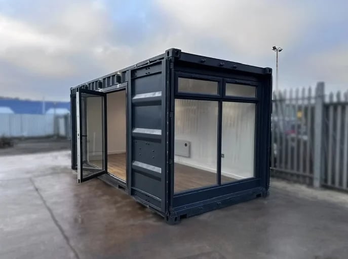 Modern black shipping container structure with glass doors and windows, positioned outdoors in an industrial yard setting