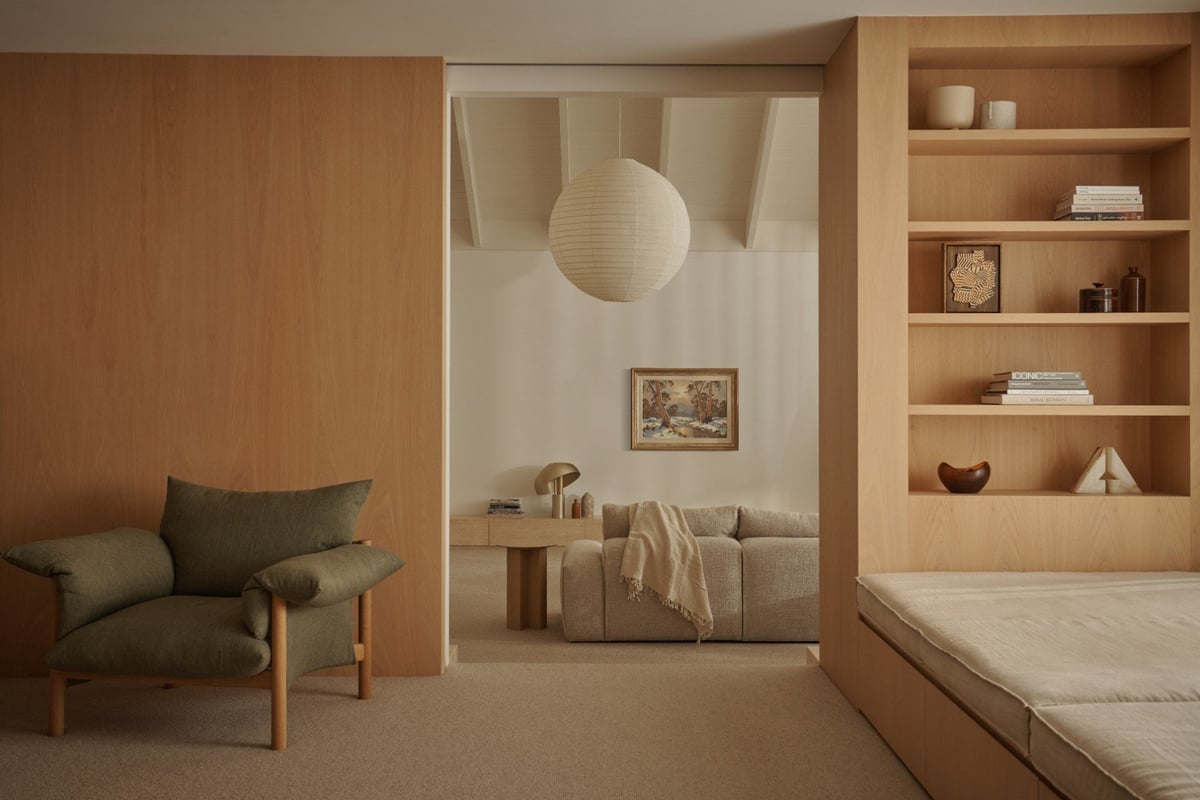 Minimalist bedroom with wooden furnishings, olive green armchair, built-in shelving, and a connected seating area with a hanging light fixture