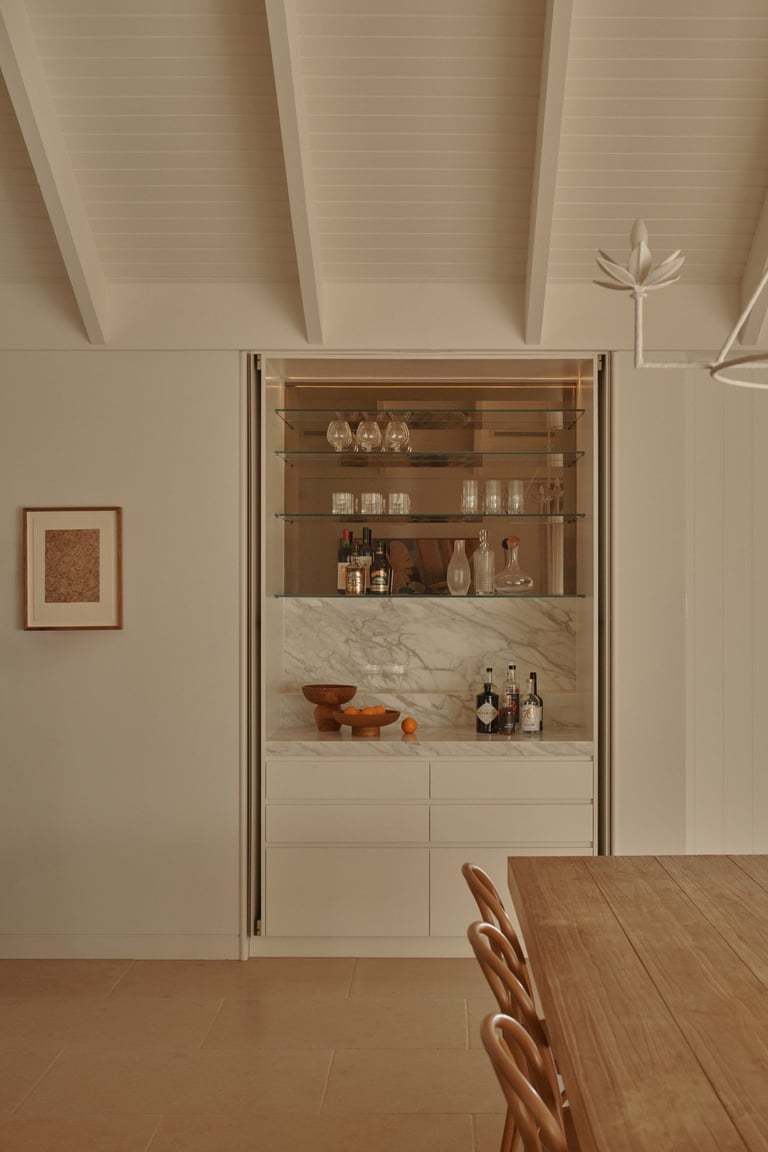 Modern kitchen with white cabinetry, open glass shelving displaying glassware and bottles, marble countertop, wooden dining table with woven chairs, and exposed white ceiling beams