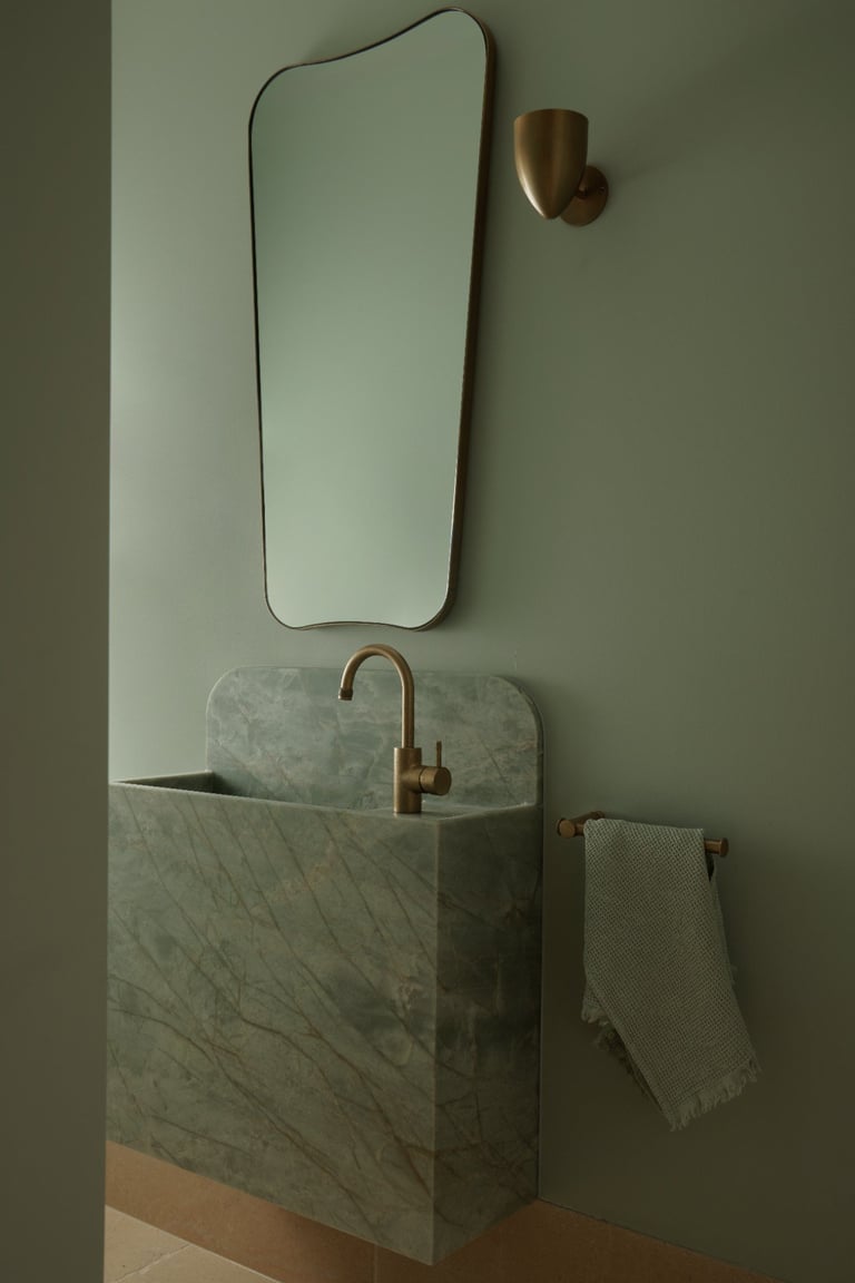 Modern bathroom sink in marble with bronze faucet, rounded mirror above, wall sconce, and towel rack against sage green wall