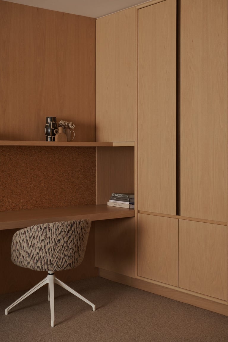 Minimalist home office with warm wood cabinetry, built-in desk, cork backing, potted plant, and upholstered chair with white legs