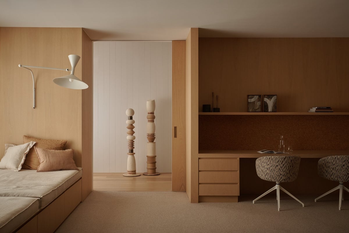 Modern minimalist bedroom with wooden built-in bed on left, stacked sculptural objects in center, and work desk with chair on right, lit by pendant lamp