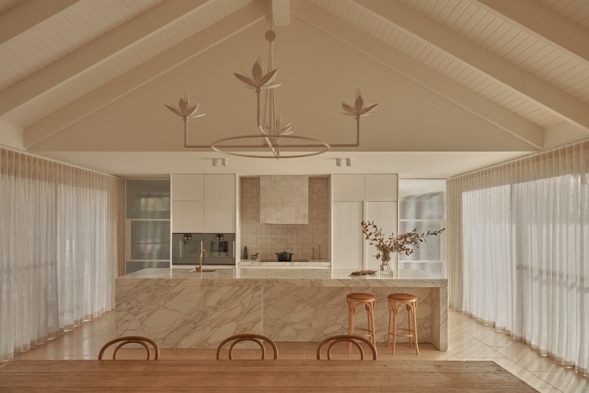 Modern kitchen and dining space with cream cabinetry, wooden beams, pendant lighting, and sheer curtains flanking glass doors