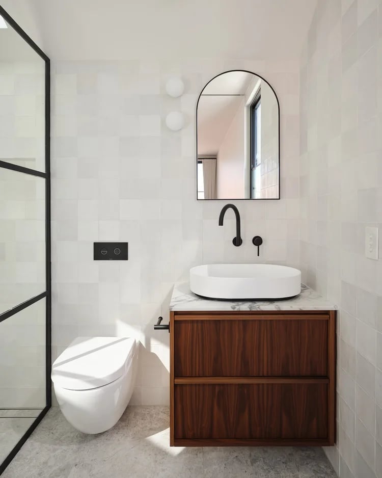 Modern bathroom with white walls, arched mirror, wooden vanity with white vessel sink, black fixtures, and walk-in shower with grid glass door