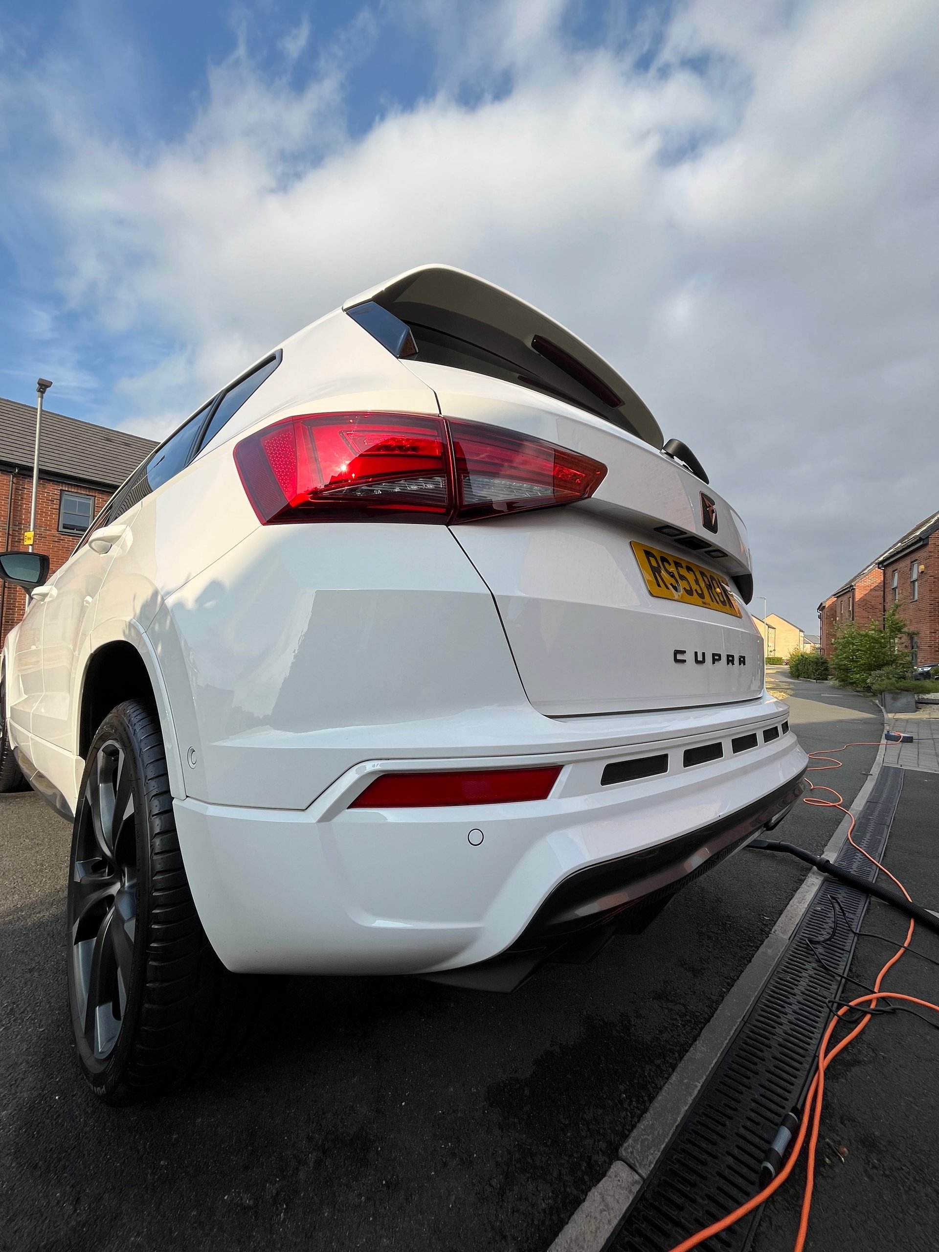 White Cupra car parked on street with red taillights visible, charging cable attached