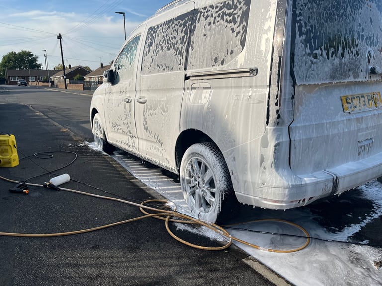White van being washed with a hose at a car wash facility, with soapy water and cleaning equipment visible