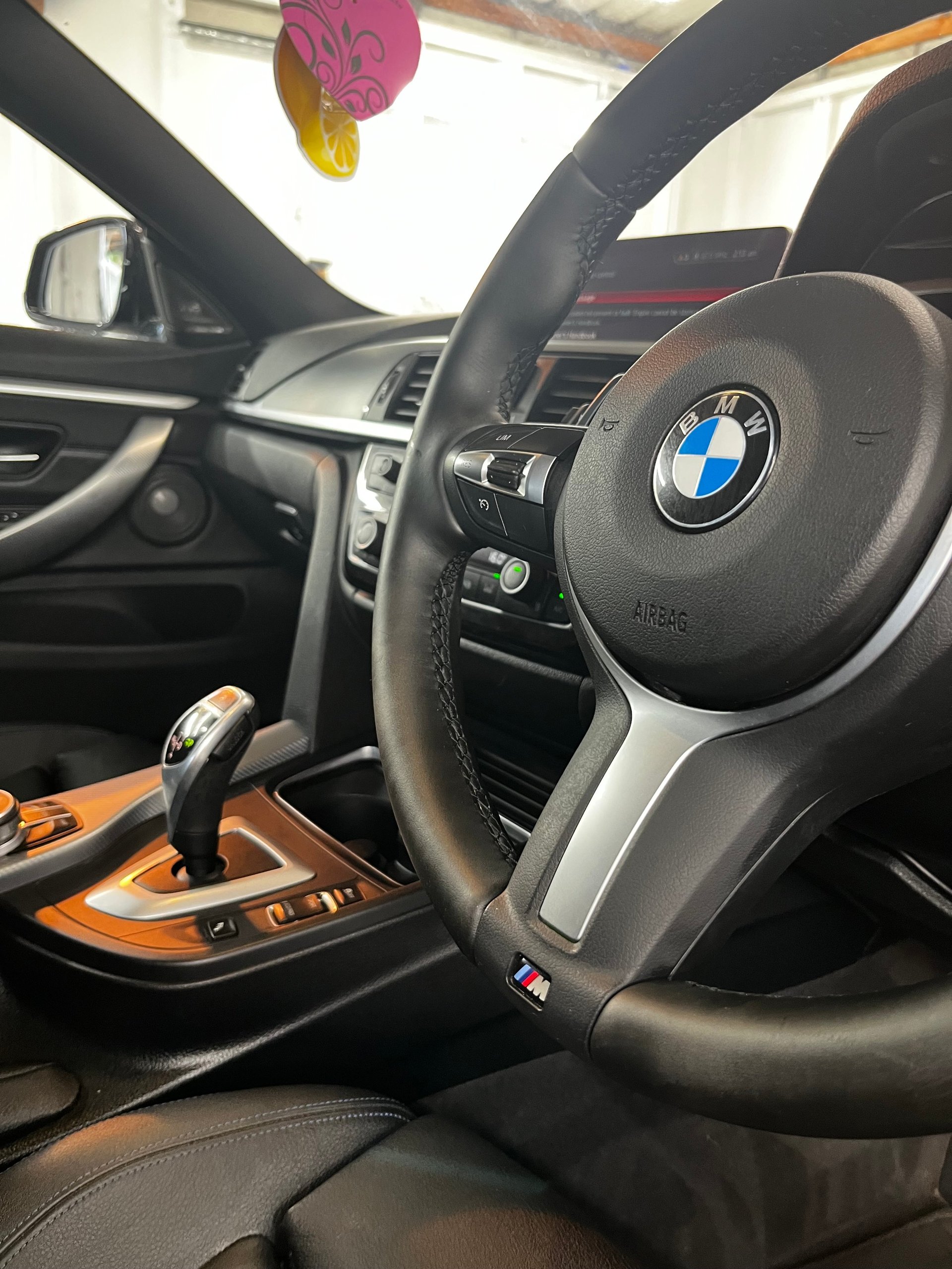 Interior view of a BMW car showing the steering wheel with logo, black and wood-grain dashboard, and pink air freshener hanging from the rearview mirror