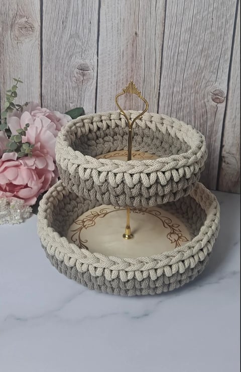 Two-tiered cream crocheted tiered stand with gold handles holding pink flowers and decorative items on white marble surface