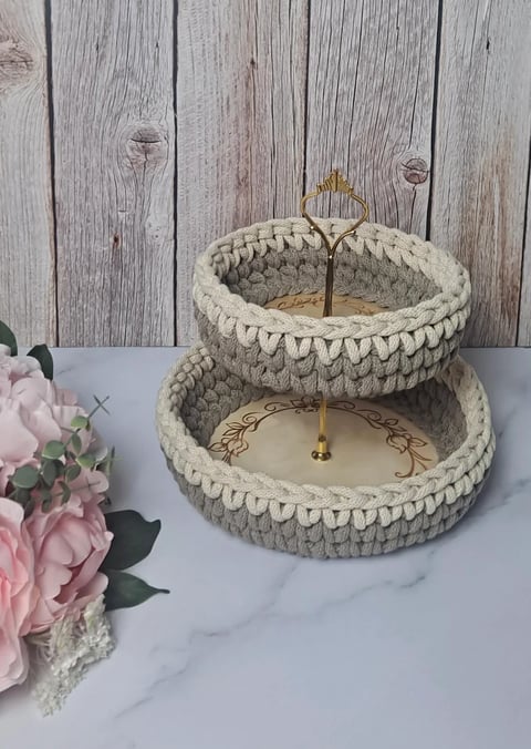 Two-tiered cream crochet basket with brass stand, decorated with pink flowers on marble surface against wooden background