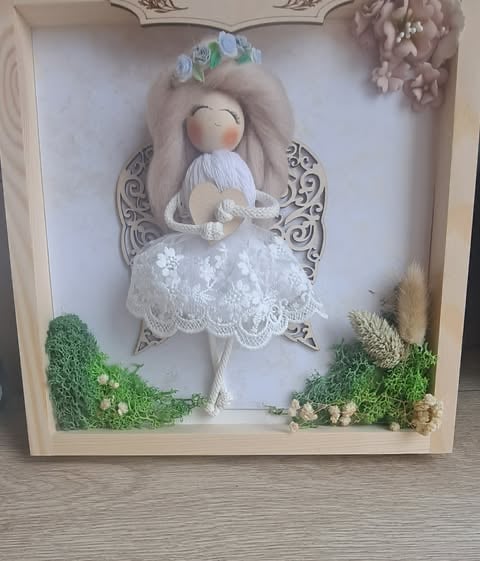 Decorative fabric angel doll in white lace dress with gray hair and wings, displayed in a shadow box with moss and flowers