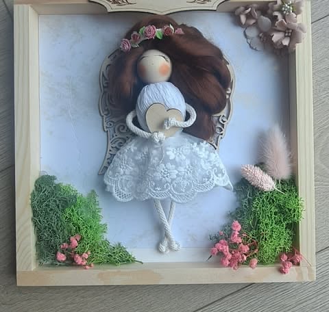 Handmade doll in shadow box with brown hair, white lace dress, and blue bonnet, surrounded by moss and pink flowers