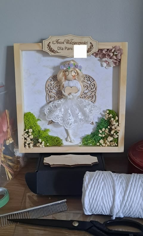 Framed decorative angel figurine with white lace wings, surrounded by moss and white flowers in wooden frame on display