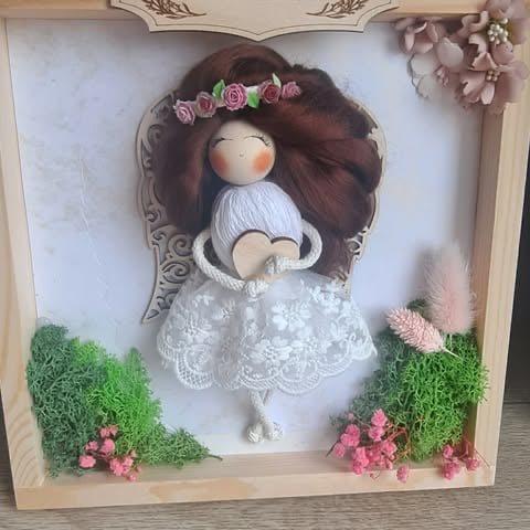 Decorative doll with brown hair, flower crown, and white lace dress displayed in a framed shadow box with moss, pink flowers, and ornamental details